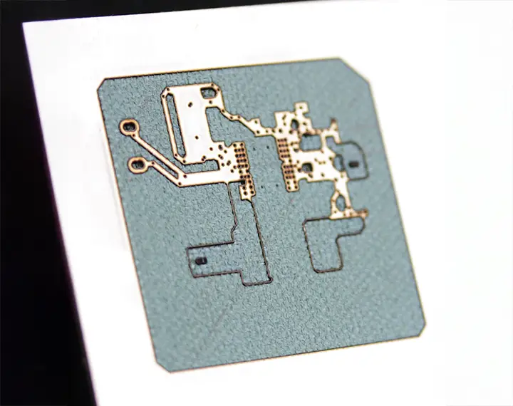 Gros plan sur un circuit imprimé, présentant des traces conductrices métalliques et des composants sur un substrat carré et grisâtre. La précision d'un laser marqueur assure une gravure détaillée, le tout affiché sur un fond blanc.