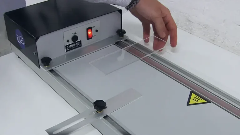 Une personne place une feuille d'acrylique transparent sur une machine de table de l'atelier Machines, dotée d'un interrupteur et d'un voyant lumineux, probablement utilisée pour découper ou plier des matériaux plastiques.
