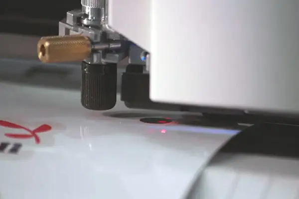 Gros plan d'un plotter de découpe en fonctionnement, avec une lame tranchante et des boutons de réglage découpant un dessin sur une feuille de vinyle blanc.
