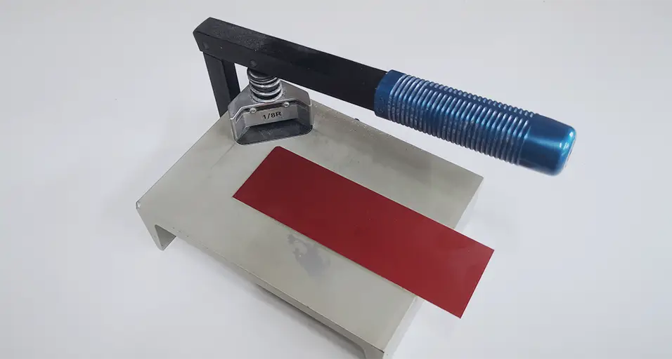 Un outil de presse en métal avec une poignée bleue est monté sur une base argentée, maintenant une feuille rectangulaire rouge sur une surface blanche - idéal pour une utilisation dans n'importe quel atelier de machines.