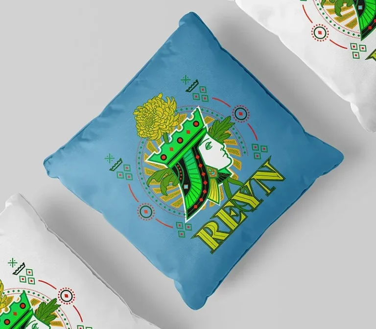 Un coussin bleu créé avec l'impression DTF représente un visage de reine de profil portant une couronne verte, des motifs géométriques et une fleur jaune, avec le mot REIGN en dessous. Deux coussins blancs avec des motifs partiels se trouvent à côté.