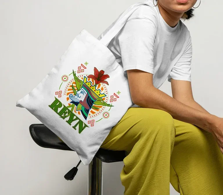 Une personne vêtue d'un t-shirt blanc et d'un pantalon jaune est assise sur un tabouret et tient un sac fourre-tout DTF imprimé d'un poisson coloré, d'un motif géométrique et du mot REYN.