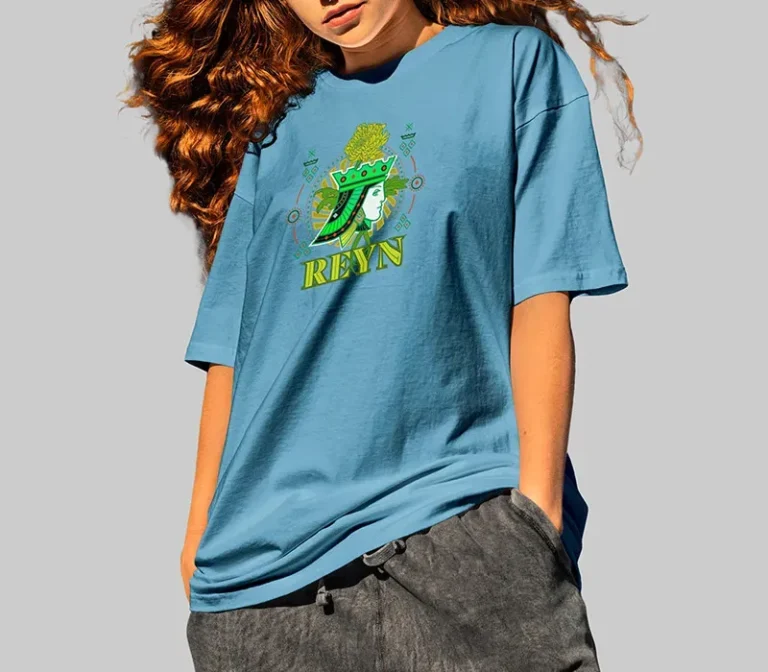 Une personne aux longs cheveux roux ondulés porte un t-shirt bleu ample avec un graphisme vert et le mot REYN, créé à l'aide de l'impression DTF, ainsi qu'un pantalon gris foncé, sur un fond gris uni.