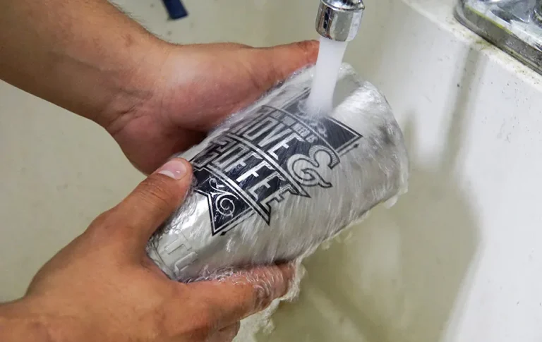 Une personne tient un mug de voyage en argent sous un robinet en train de couler, le rinçant avec de l'eau. Le mug comporte un texte et des graphiques en noir et blanc créés par un marqueur laser. L'évier et le robinet sont blancs.