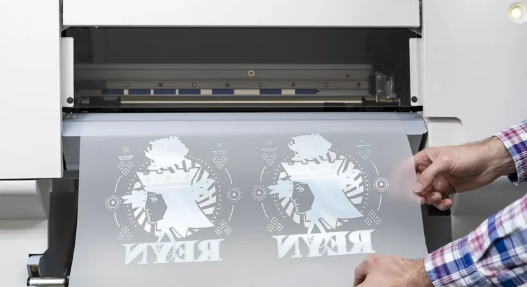 Une personne tire d'une machine à imprimer DTF à impression blanche une feuille contenant deux motifs imprimés en miroir, chacun représentant une silhouette stylisée d'un visage de femme et le mot REY en lettres capitales.