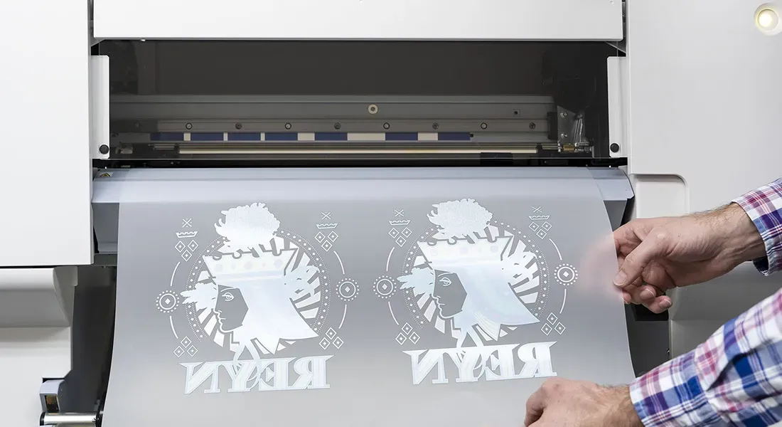 Une personne tire d'une machine à imprimer DTF à impression blanche une feuille contenant deux motifs imprimés en miroir, chacun représentant une silhouette stylisée d'un visage de femme et le mot REY en lettres capitales.
