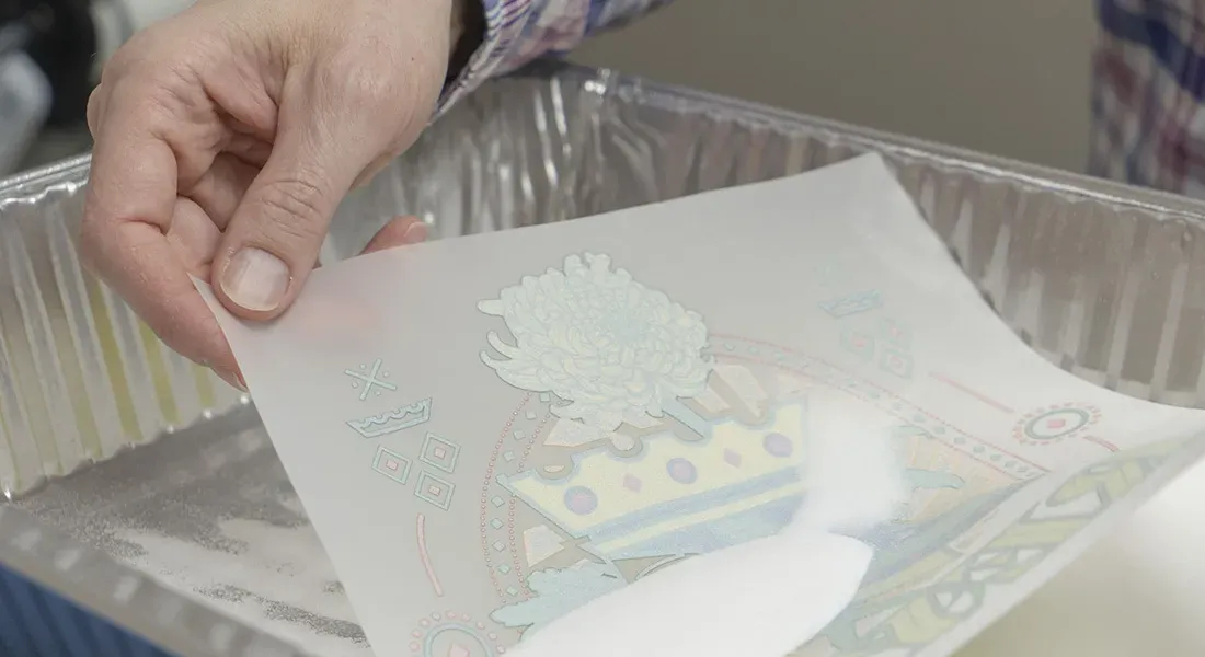 Une personne tient une feuille translucide avec une impression dtf au-dessus d'un plateau métallique rempli de poudre blanche, se préparant peut-être à un transfert ou à un processus artisanal.