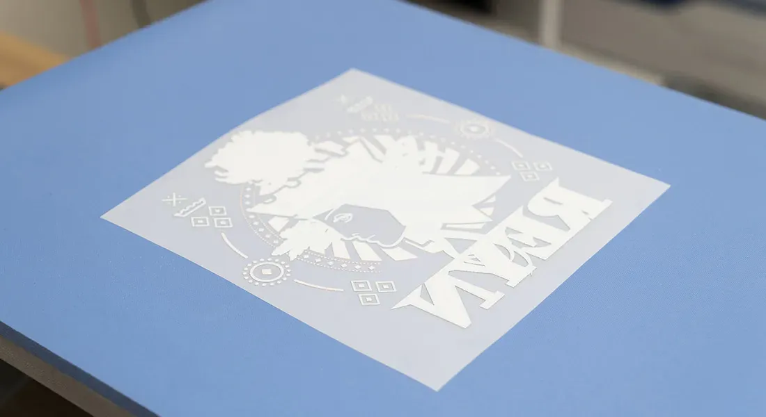 Un pochoir blanc avec un motif décoratif et un texte en miroir est placé sur une surface plane bleue, prêt à être utilisé dans le cadre d'un processus d'impression DTF ou d'artisanat.