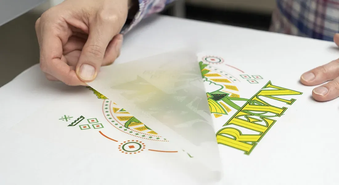 Les mains d'une personne décollent une feuille de transfert translucide d'une surface blanche, révélant un motif imprimé coloré avec des motifs géométriques et le mot "REYN", créé à l'aide de la technologie impression dtf.