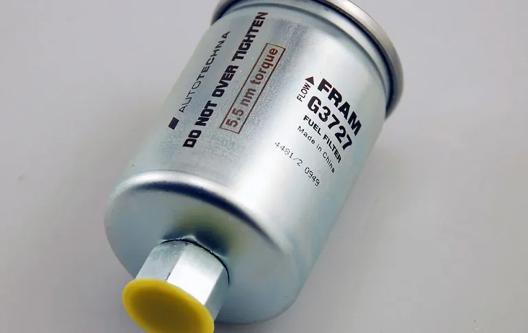 Un filtre à carburant cylindrique en métal étiqueté "FRAM G3721" est couché sur le côté, avec un texte clair marqué au laser CO2 : "NE PAS TROP SERRER", "Couple de 5,5 Nm" et "FILTRE À CARBURANT". L'extrémité du connecteur est munie d'un capuchon jaune.