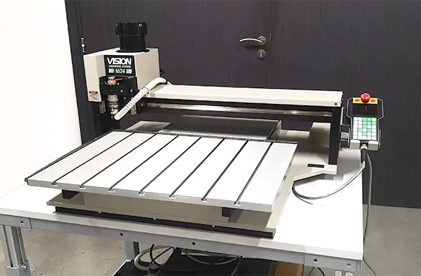 Une fraiseuse CNC de bureau avec une table de travail métallique et un panneau de commande est placée sur une table blanche devant une porte sombre fermée. La machine est étiquetée VISION et 1624R.