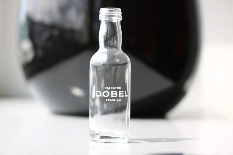 Une petite bouteille en verre transparent étiquetée Maestro Dobel Tequila se tient debout sur une surface blanche avec un arrière-plan flou et sombre. La bouteille est vide et n'a pas de bouchon.