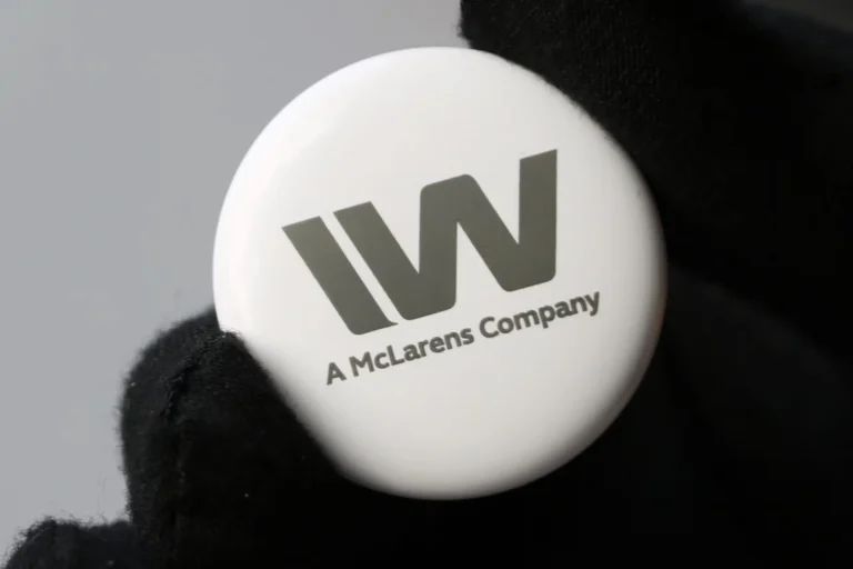 Une main gantée tient un bouton rond blanc avec un grand W stylisé et le texte "A McLarens Company" imprimé en gris, mettant en valeur la précision du marquage industriel.