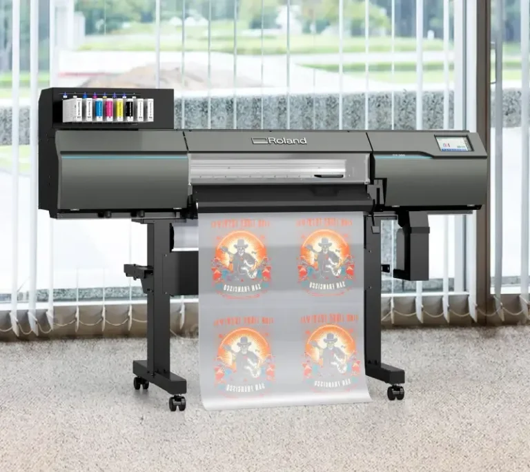 Une grande imprimante Roland se trouve devant de hautes fenêtres et imprime un rouleau d'affiches dtf d'impression représentant un personnage sur une moto avec des graphiques orange et jaune vifs et le texte "APOCALYPSE NOW". Des cartouches d'encre sont visibles sur le côté gauche.