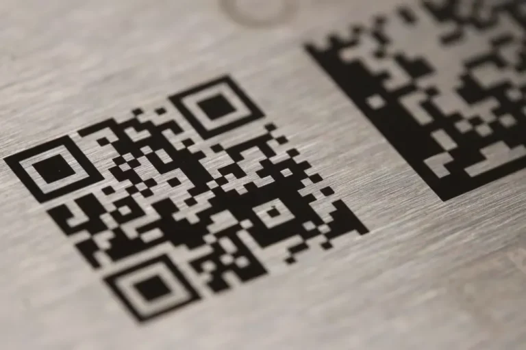 Vue rapprochée d'un code QR noir et blanc gravé au laser marqueur sur une surface métallique brossée. Un autre code QR partiel, créé à l'aide de la technologie laser co2, est visible dans le coin supérieur droit de l'image.