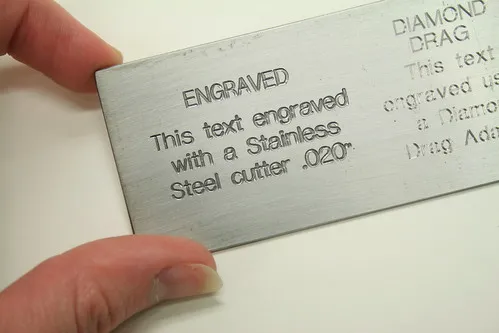 Une main tient une plaque de métal gravée du texte : "ENGRAVED Ce texte est gravé à l'aide d'une fraise en acier inoxydable .020-", mettant en évidence la précision des machines de fraisage héliogravure. La plaque repose sur une surface blanche.