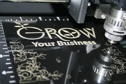 Gros plan sur une machine à laser CO2, présentant la technologie marquage, qui grave la phrase Grow Your Business et des motifs décoratifs sur une surface noire, avec des repères de mesure visibles sur le côté.