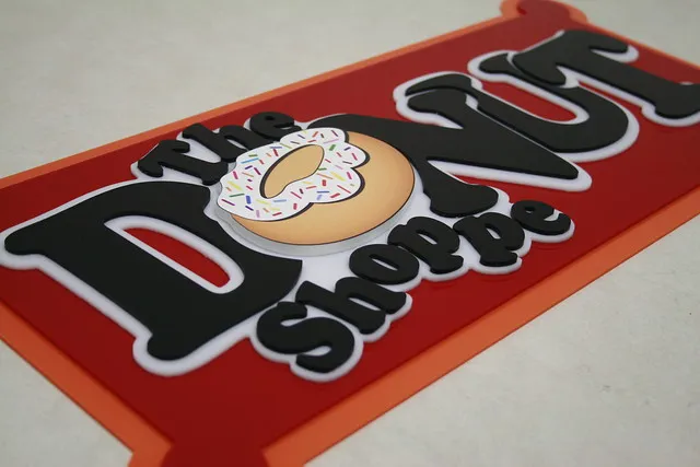 Une enseigne indique The Donut Shoppe avec un donut illustré comme la lettre O de Donut. Le beignet, recouvert d'un glaçage blanc et de paillettes colorées, suggère la précision des machines de fraisage en héliogravure. L'enseigne a un fond rouge et une bordure orange.