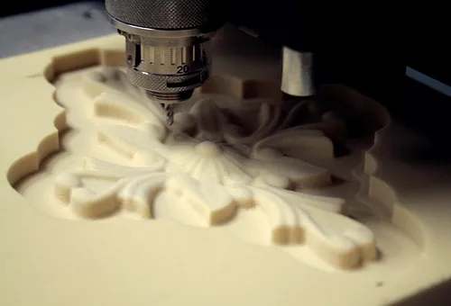 Une machine CNC grave un motif floral complexe dans un matériau de couleur claire. L'outil de gravure de la machine est positionné au-dessus de la pièce, façonnant des motifs détaillés et symétriques.