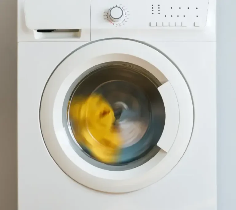 Vue de face d'une machine à laver blanche avec des vêtements qui tournent à l'intérieur, y compris un vêtement jaune visible, pendant un cycle de lavage - parfait pour nettoyer les articles avant d'appliquer des impressions dtf. Le bouton et le panneau de commande se trouvent en haut de la machine.