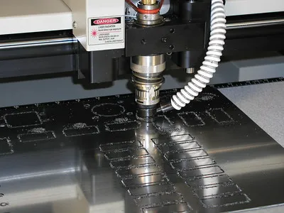 Une machine CNC découpe des formes et des motifs précis dans une feuille de métal, présentant des machines avancées de fraisage en héliogravure. La tête de coupe active produit des copeaux de métal autour des zones coupées, et une étiquette d'avertissement est visible près de la tête de l'outil.