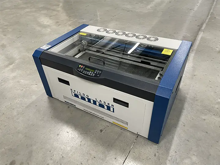 Une machine d'occasion laser CO2 Epilog bleue et blanche est posée sur un sol en béton nu dans un grand espace intérieur vide.