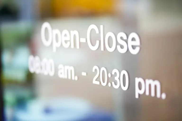 Une porte en verre affiche un texte blanc indiquant les heures d'ouverture, avec la mention Open-Close 09:00 am - 20:30 pm, probablement créé à l'aide d'un traceur de découpe. L'arrière-plan est flou et mal cadré.