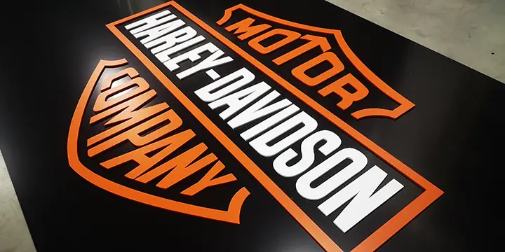 Logo Harley-Davidson comportant les mots Motor and Company en orange et Harley-Davidson en blanc, sur fond noir. Le texte est disposé à l'intérieur de deux boucliers aux contours orange, idéaux pour un travail de précision avec un laser découpeur.
