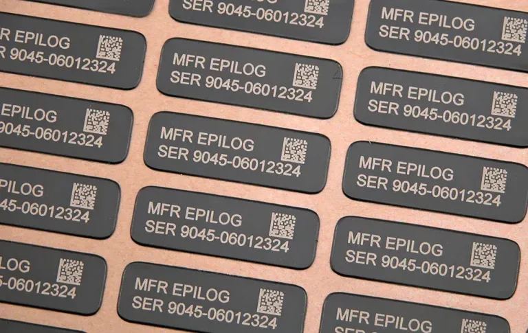 Vue rapprochée de plusieurs étiquettes rectangulaires noires créées à l'aide d'un laser graveur, comportant un texte imprimé en blanc indiquant MFR EPILOG SER 9045-06012324 et un code QR sur chacune d'entre elles, disposées en rangées ordonnées sur une surface plane.
