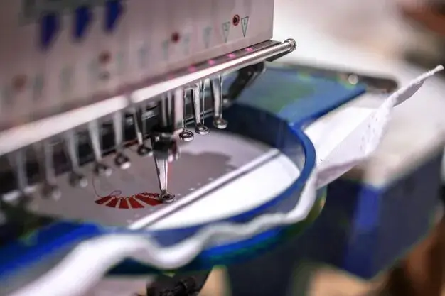 Vue rapprochée d'une machine à broder cousant un motif rouge et blanc sur un morceau de tissu blanc maintenu dans un cercle de broderie bleu.