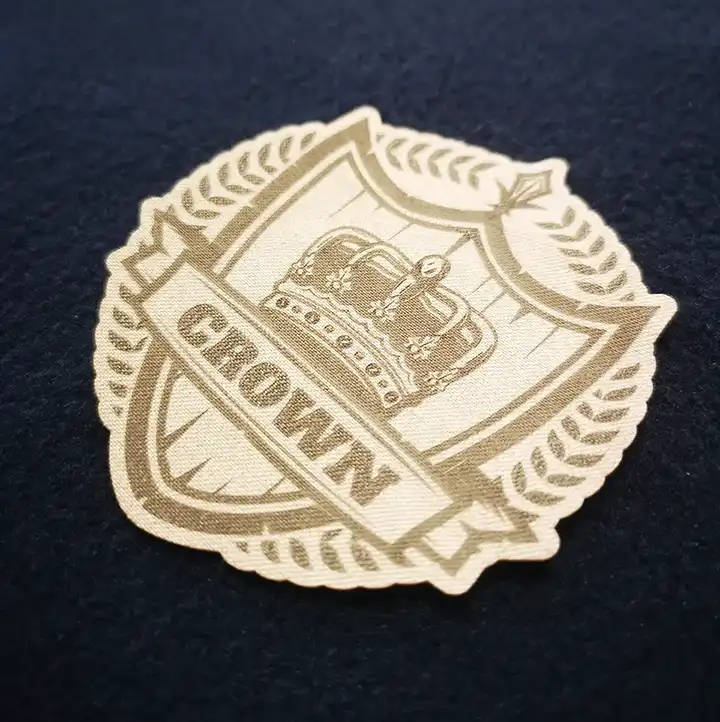 Un écusson en tissu représente une couronne à l'intérieur d'un écusson entouré de feuilles de laurier ; le mot "CROWN" apparaît sur une bannière sous la couronne, et l'écusson est placé sur une surface sombre et texturée.