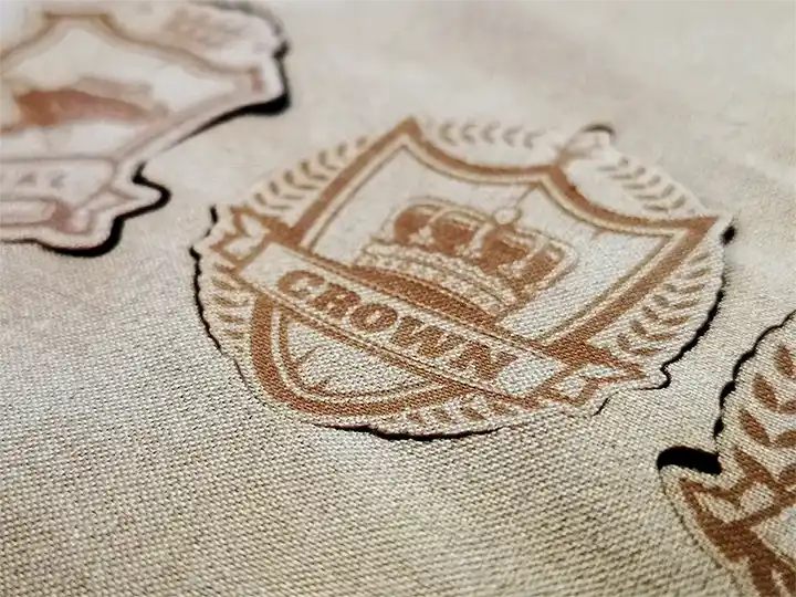 Gros plan d'un tissu beige avec un écusson brodé représentant une couronne et le mot CROWN en dessous, entouré d'une couronne de laurier. D'autres écussons similaires sont partiellement visibles à l'arrière-plan.