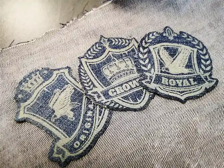 Trois écussons en tissu brodé avec des motifs de bouclier sont cousus sur du denim. Les écussons comportent des symboles tels qu'une couronne, un oiseau, des couronnes de laurier et un texte indiquant ROYAL, CROWN et EST.