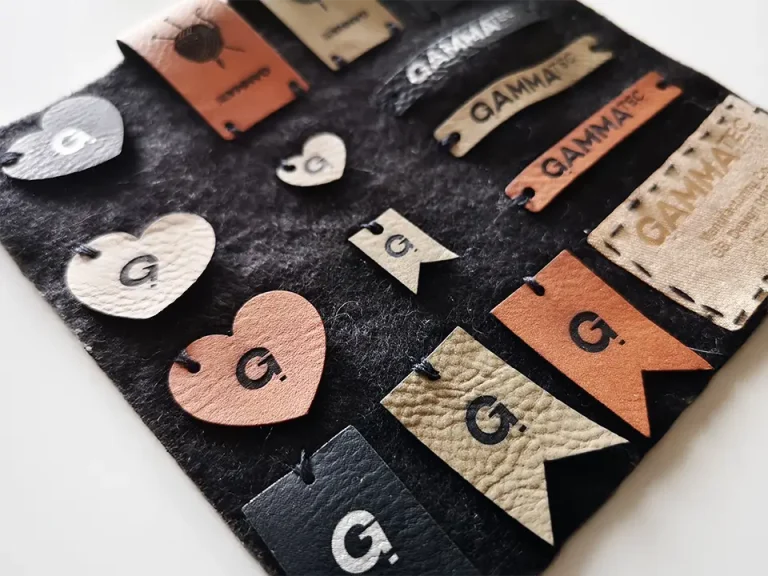 Une collection d'étiquettes en cuir et en tissu, comprenant des cœurs, des rectangles et des drapeaux, tous attachés à un fond de feutre noir. La plupart des étiquettes comportent la lettre G. et le mot GAMMA dans différentes polices.