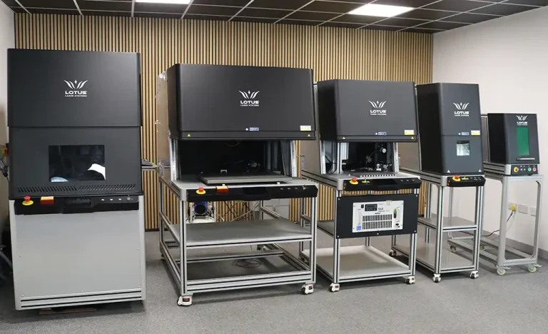 Quatre grandes machines de gravure laser industrielle GAMMA-TEC de couleur noire, montées sur des châssis métalliques, sont alignées contre un mur recouvert de panneaux de bois verticaux dans une pièce propre et moderne au plafond carrelé.