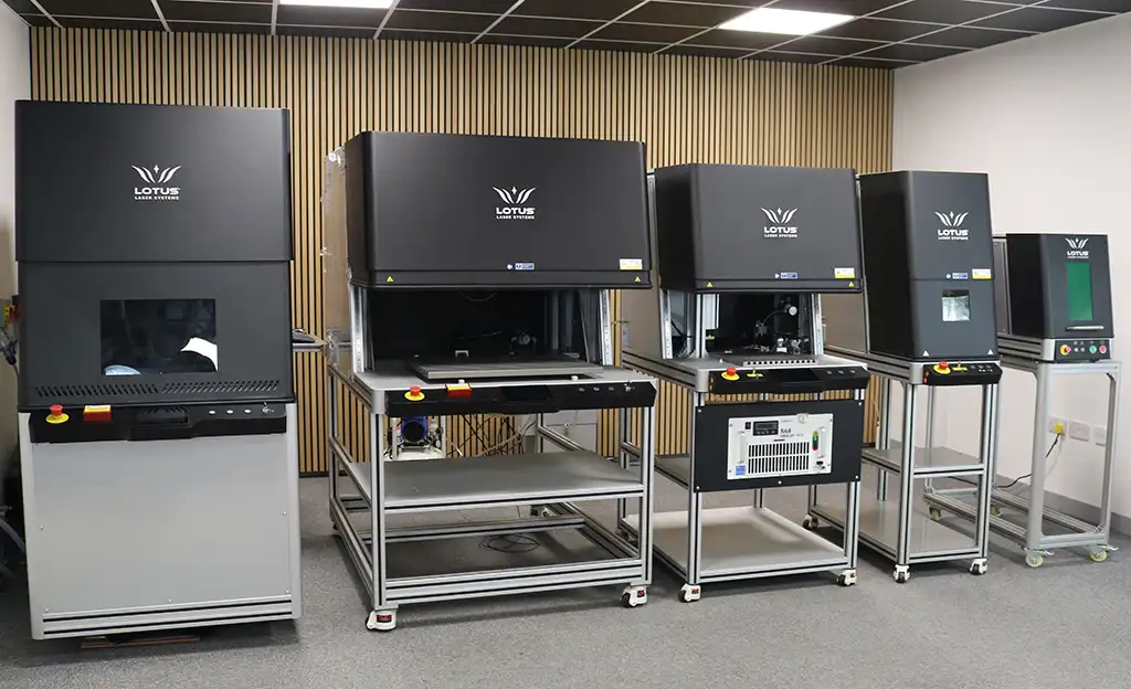 Quatre grandes machines de gravure laser industrielle GAMMA-TEC de couleur noire, montées sur des châssis métalliques, sont alignées contre un mur recouvert de panneaux de bois verticaux dans une pièce propre et moderne au plafond carrelé.