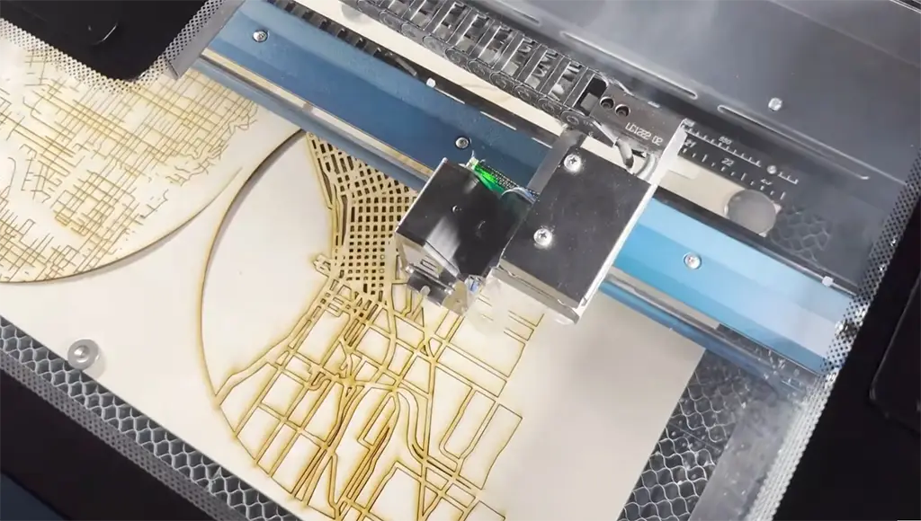 Une machine de découpe laser CO2 Epilog travaille sur un morceau de bois clair, découpant une carte détaillée avec des rues et des intersections.