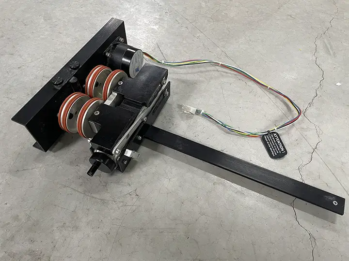 Un accessoire rotatif pour machine Epilog doté de roues cerclées de rouge, d'un moteur, de câbles et d'un long bras métallique noir est posé sur un sol en béton gris.