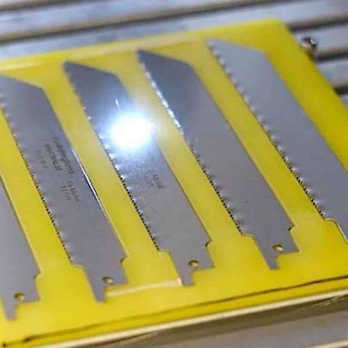 Cinq lames de scie en métal sont disposées dans des fentes parallèles sur un support en plastique jaune, qui est placé sur une surface métallique avec des rainures horizontales. La lumière se reflète sur la lame centrale.