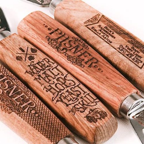 Quatre décapsuleurs à manche en bois sont disposés en ligne sur une surface blanche. Chaque manche est gravé de textes et de motifs décoratifs, créés à l'aide d'une machine laser précise, dont le mot Pilsner.