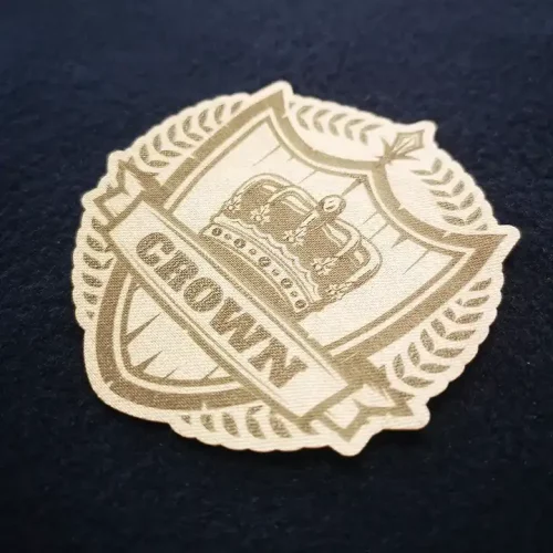 Un écusson en tissu représente une couronne à l'intérieur d'un écusson entouré de feuilles de laurier ; le mot "CROWN" apparaît sur une bannière sous la couronne, et l'écusson est placé sur une surface sombre et texturée.