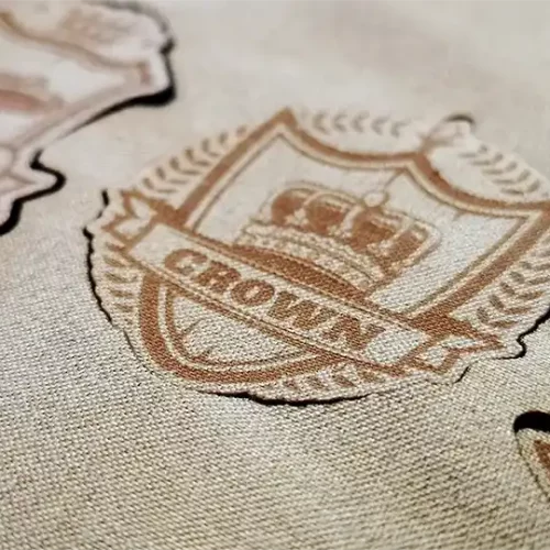 Gros plan d'un tissu beige avec un écusson brodé représentant une couronne et le mot CROWN en dessous, entouré d'une couronne de laurier. D'autres écussons similaires sont partiellement visibles à l'arrière-plan.