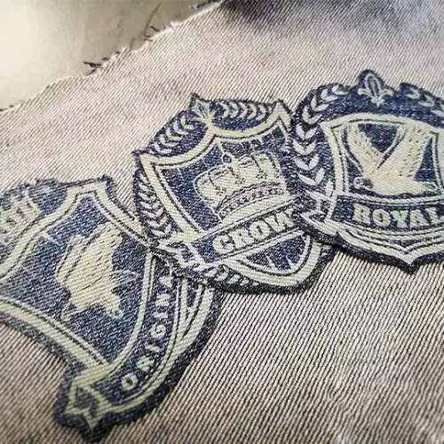 Trois écussons en tissu brodé avec des motifs de bouclier sont cousus sur du denim. Les écussons comportent des symboles tels qu'une couronne, un oiseau, des couronnes de laurier et un texte indiquant ROYAL, CROWN et EST.