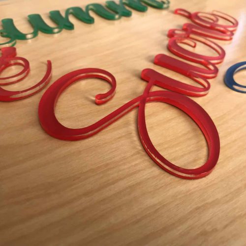Gros plan sur des lettres acryliques cursives et colorées, notamment vertes, rouges et bleues, disposées sur une surface en bois clair. Les lettres brillantes, parfaites pour tout fournisseur de matière à graver, reflètent magnifiquement la lumière.