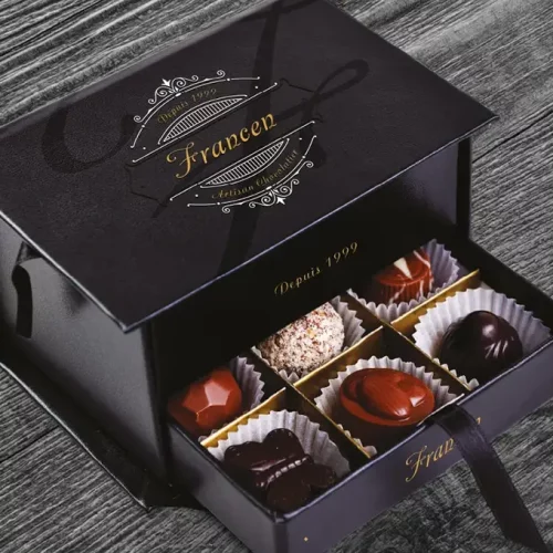 Une boîte noire avec des accents dorés et "Francen" créé par l'imprimante sur objet contient six chocolats assortis dans des gobelets en papier individuels, élégamment présentés dans un tiroir coulissant sur une surface en bois.