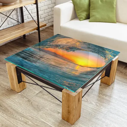 Table basse carrée avec un plateau en verre, comportant une image imprimée sur objet d'une vague au coucher du soleil. La table a d'épais pieds en bois, des supports en métal noir, et repose sur un plancher en bois près d'un canapé blanc avec des coussins verts.