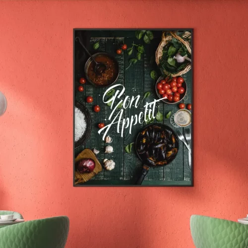Une image encadrée sur un mur rouge - créée avec l'imprimante sur objet - montre des ingrédients et des plats sur une surface en bois vert foncé avec le texte Bon Appétit en écriture blanche. Deux chaises vertes et des lampes suspendues blanches sont partiellement visibles.