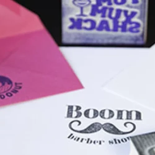 Une collection de timbres en bois, fabriqués avec une précision proche du travail à la machine, et des enveloppes sur une surface sombre. Les timbres présentent des motifs tels que « MUY MUY SHACK » et « Boom barber shop » avec une moustache. Les enveloppes en dessous affichent fièrement des empreintes en violet et bleu.