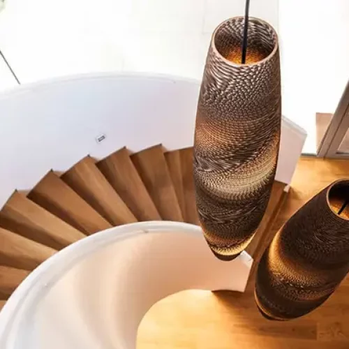 Un escalier en colimaçon avec des marches en bois mène à un espace lumineux. Deux grandes suspensions au design complexe sont suspendues au-dessus, projetant une lumière chaleureuse sur les escaliers. De grandes fenêtres offrent une vue sur lextérieur, ajoutant une lumière naturelle à lespace qui semble aussi inspirante que les créations de Think Paper.