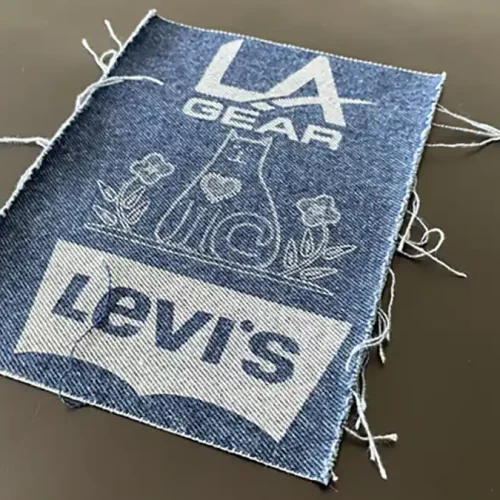 Un patch en denim parfait pour la personnalisation textile, avec « LA Gear » en haut, une forme géométrique avec des fleurs au centre et « Levis » en bas. Les bords effilochés ajoutent du caractère à sa surface unie.